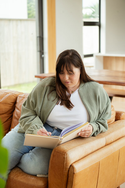 Chantal Hewitt writing in a notebook wearing a white shirt and green jacket while sitting on a leather tan couch - Family Autism Support Coach at Chantal Hewitt Coaching5