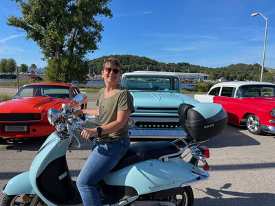 Amy Gould riding a blue moped, parked near classic cars on a sunny day
