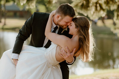 A groom in a black suit joyfully lifts his bride in a white dress near a serene pond. They touch foreheads, smiling, surrounded by soft greenery.