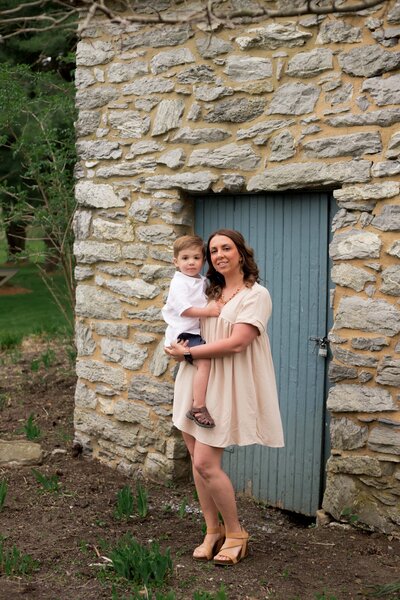 Mom in a cream dress holding young son in front of a stone building