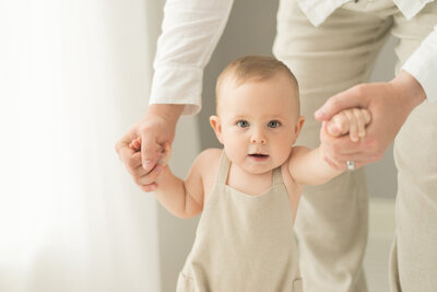 Baby in neutral overalls takes small steps while holding a parent's hands. A sweet milestone session by Moments in Light Photography.