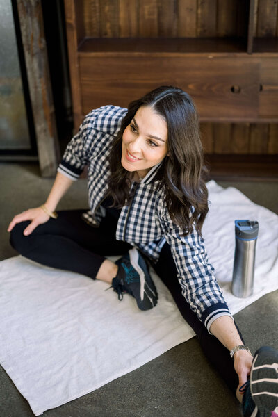 Woman in a plaid shirt sitting on the floor performing a hamstring stretch.