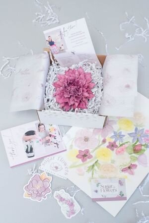 a shipping box with a purple dahlia sugar flower surrounded by shipping material like paper shred, tissue paper, thank you cards, business cards, stickers, and design sketches from sugar flowers by kelsie cakes