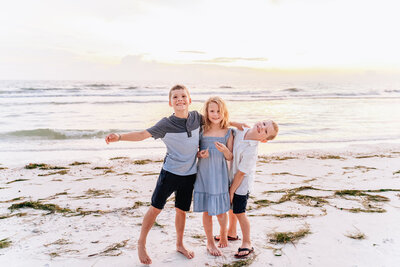 Three children stand on a sandy beach at sunset, playfully posing and smiling for the camera with the ocean in the background.
