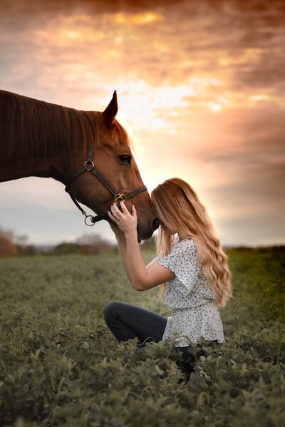 Girl sitting in field with head on horses nose during sunset