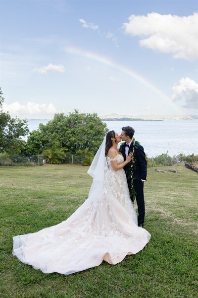A newlywed couple kissing in a field with a rainbow behind them 