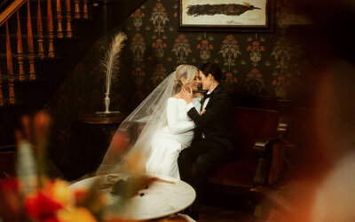 Bride and groom sitting together indoors, sharing an intimate embrace in classic wedding attire.