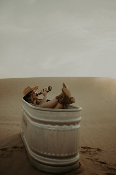 Portrait of a woman in a drinking trough wearing cowboy hat and boots  while drinking from a bottle of champagne.