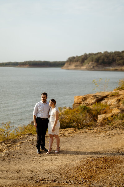 A couple stands on the red rocky terrain of Rockledge Park in Texas, dressed in timeless formal wear as they look out over the lake. Her white lace dress and strappy heels complement his classic black-and-white attire, creating a romantic and elegant moment against the natural backdrop.
