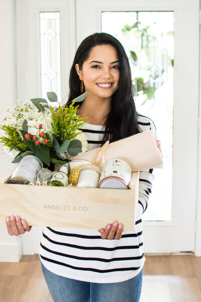 Smiling woman with dark hair standing and holding a wooden box filled with gifts
