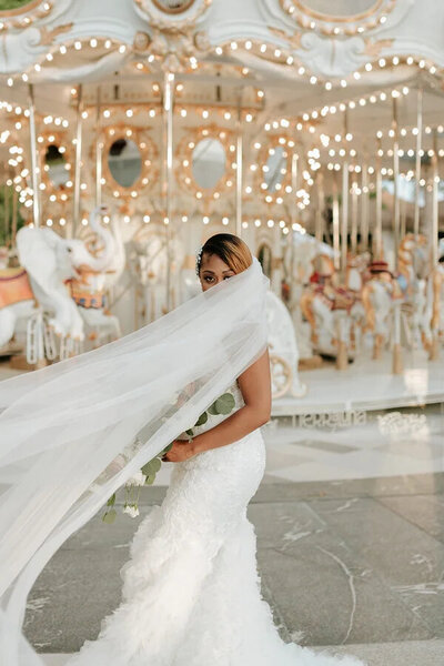 Bride stands in front of carousel with veil covering her face
