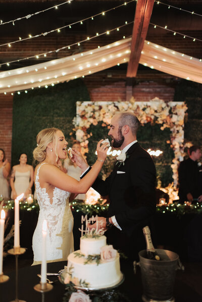 Bride and groom cut the cake and feed each other at their reception at Distillery 244 Old Town while surrounded by the most beautiful lighting provided by LeLuci Lighting in Wichita Kansas