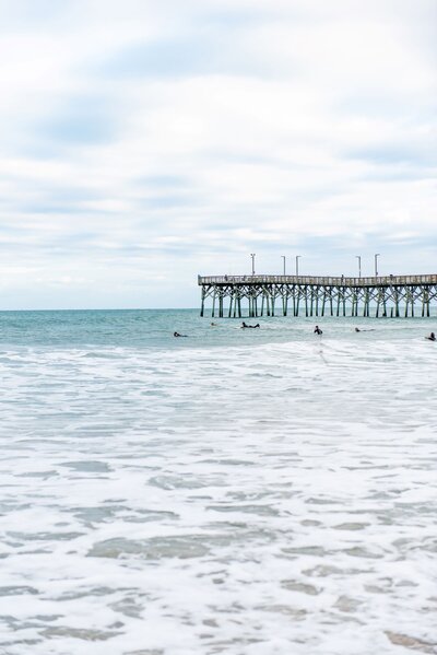 a cloudy sky covers the blue ocean full of surfers with the surf city, north carolina pier in the distance