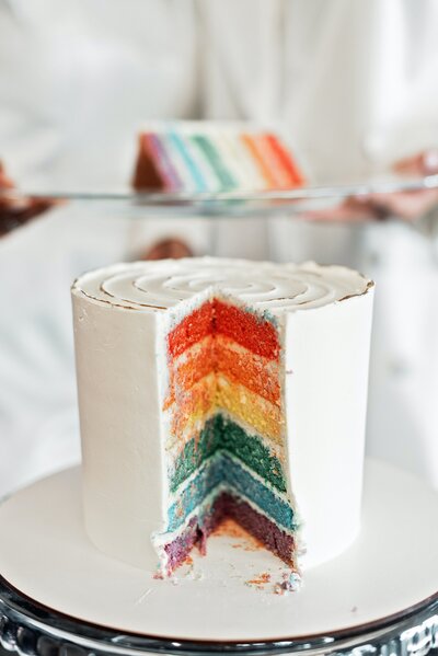 A tall white cake with a piece cut out showing a rainbow interior.