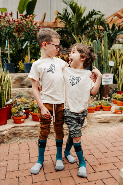 Two boys make silly faces at each other inside greenhouse surrounded by plants.