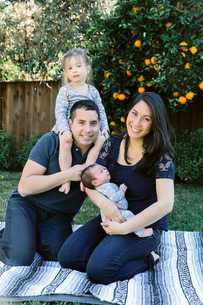 Dad sits with daughter on his shoulders while mom holds baby all smiling at the camera