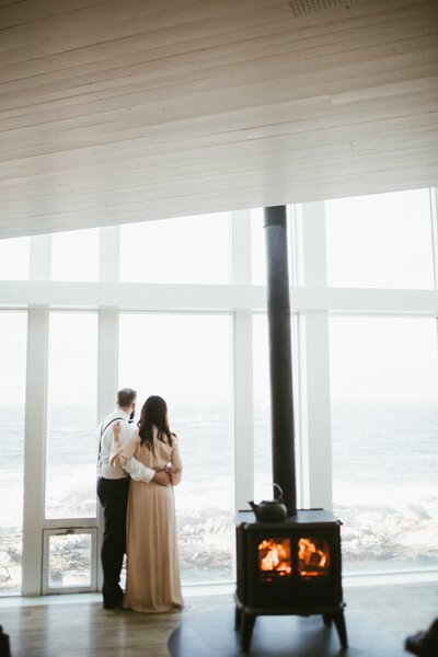 bride and groom look out the window beside a lit woodstove