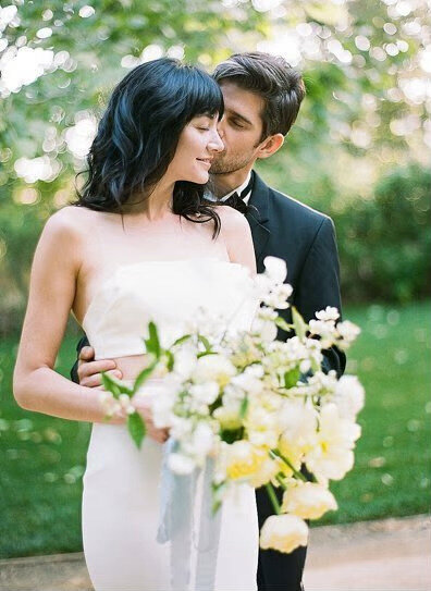 Kohnur modern romantic couple with bride in natural glowing makeup and short hair with bangs embracing in a green garden park.
