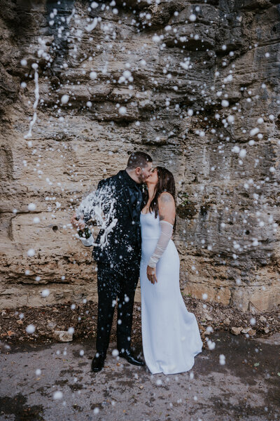 Newlyweds share a kiss as champagne sprays around them against a rocky cliff, celebrating with a playful and spontaneous moment.