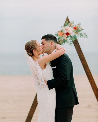 A couple kisses in front of a wooden triangle with flowers at their Cape May beach wedding.