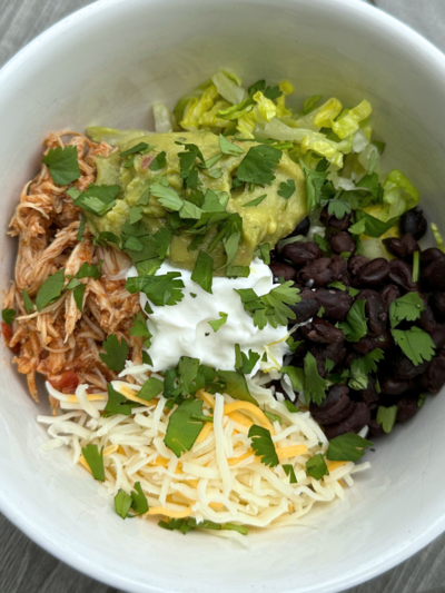 Burrito bowl with shredded chicken, black beans, lettuce, shredded cheese, sour cream, guacamole, and cilantro in a white bowl.