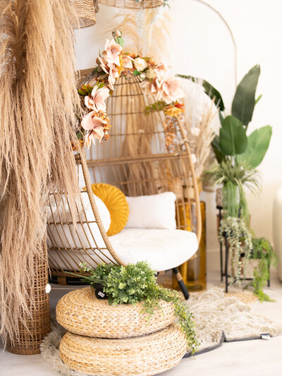 Cozy bohemian nook with a wicker egg chair adorned with pink flowers, soft cushions, and lush green plants. Warm, inviting, and peaceful ambiance.