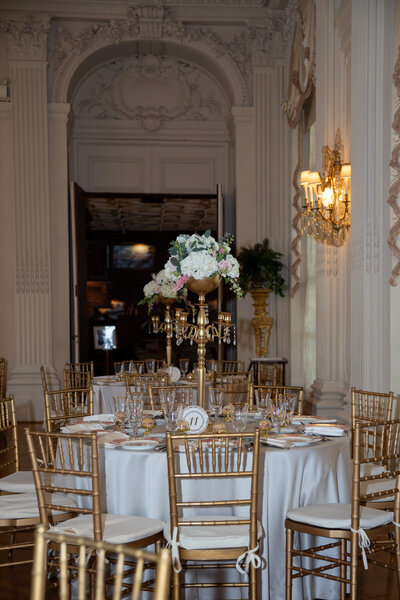 New England Wedding Photographer Pricing | Kelsey Sheehan Photography Timeless Rhode Island Weddings | Elegant dining room set for an event with a round table, white tablecloth, gold chairs, and a tall floral centerpiece. Soft lighting and ornate decor create a sophisticated tone.