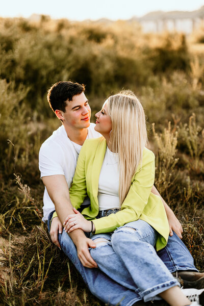 The bride to be sits between her fiancé's legs while he wraps his arms around her and they share a loving glance