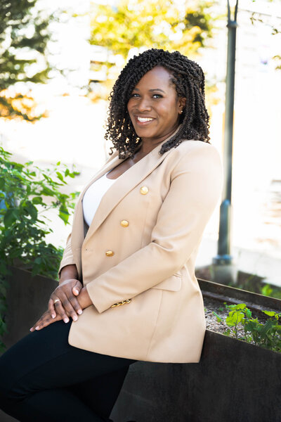 Outdoor professional portrait of a smiling woman in a beige blazer with gold buttons, leaning against a planter with greenery, set against a softly blurred background of trees and urban elements.
