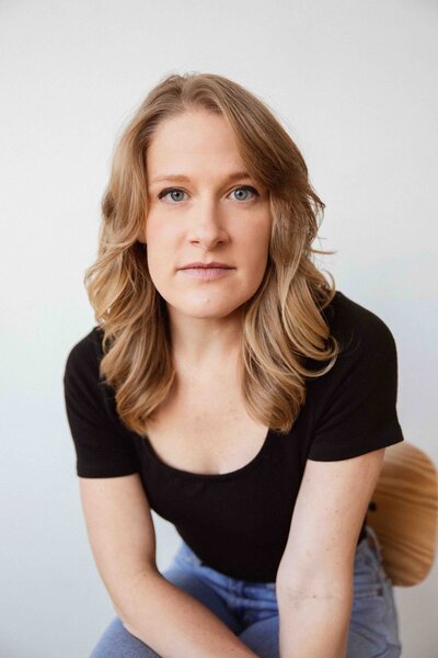 A woman with shoulder-length wavy hair and a black top sits on a wooden chair, looking directly into the camera with a calm and confident expression, framed by a clean white background.