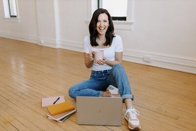 Woman with dark hair and white t-shirt and jeans sitting on floor with laptop and pink coffee mug