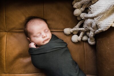 Newborn baby swaddled in a dark blanket, sleeping peacefully on a brown leather couch beside a knit throw, perfectly captured by a family photographer from Pittsburgh, PA.