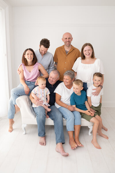 A large multi-generational family is gathered at Dandelion Studio in Moira, Northern Ireland. The group includes grandparents, parents, and young children, all posed on and around a white sofa. Everyone is smiling and laughing, capturing the joy and connection of this close-knit family in a light, airy setting.