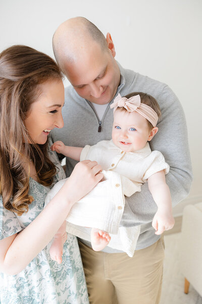 8 month old baby girl wearing pink bow smilies while being held by her parents during boston family photos