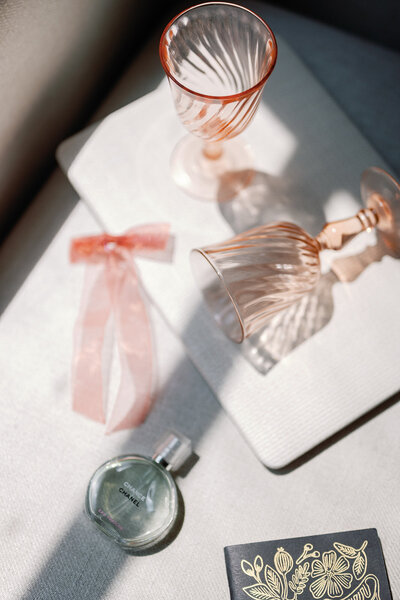 Flat lay of pink glassware, a perfume bottle, and a pink bow on a laptop