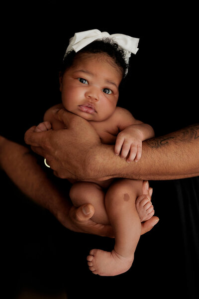 Newborn baby with a white bow on her head, held securely in the arms of an adult against a black background. The baby has a calm expression and is looking directly at the camera.