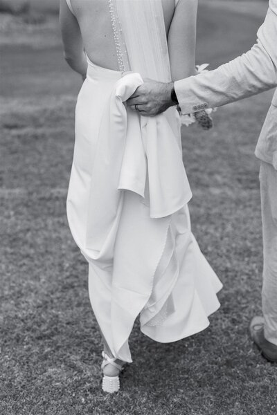 A person holding up the train of their partner's wedding dress as they walk together 