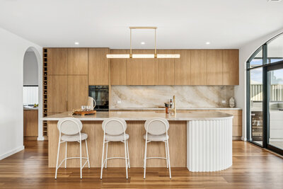 A bespoke kitchen in the Marisol home, featuring warm oak cabinetry, a fluted island bench, and stone splashback with gold accents.