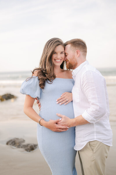 husband and wife hugging on beach, holding baby bump