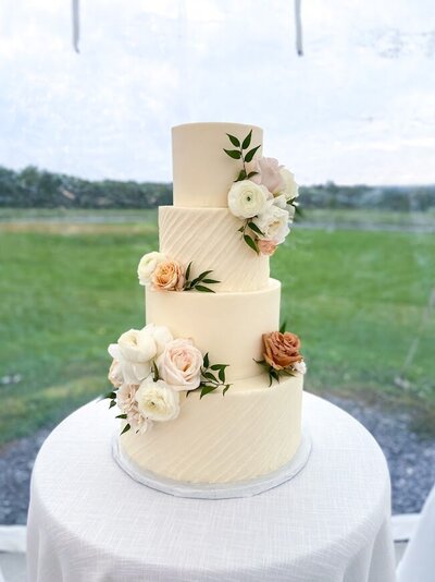 A four tier wedding cake sits on a round cocktail table with a white table cloth. there's a field of grass and line of trees blurred in the background of the cake. The cake has alternating tiers of smooth white and diagonal textured white frosting. with clusters of flowers in whites, pinks, and coffee tan.