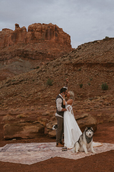 Couple having their first kiss with their dog sitting next to them. They are in their wedding attire standing on rugs with the moab desert in the background