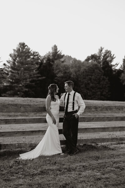 A bride and groom stand next to a wooden fence in an outdoor setting captured by a New England wedding photographer. The bride is in a long white dress, the groom wears suspenders and a tie, all set against trees. This black-and-white image epitomizes fine art weddings.