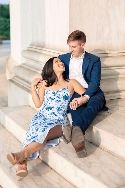 A couple sits on stone steps. The woman, in a blue floral dress, leans back smiling at the man in a blue suit. They hold hands and gaze at each other warmly against a large stone structure.