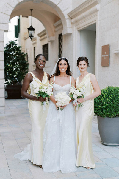 Smiling bride in white gown with two bridesmaids in champagne dresses, holding white rose bouquets in Perast