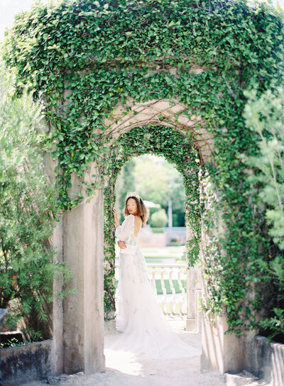 Hawaiian bridal inspiration shoot. Model is wearing a Samuelle Couture gown that is a dusty purple grey color with many layers of different lengths. She is sitting on an ivory bench. Her leg is on the bench and her mid calf to toes are showing. The layers are cascading down the floor. She has a floral side headpiece on her dark hair. Photographed for destination wedding inspiration by wedding photographers in Charleston Amy Mulder Photography