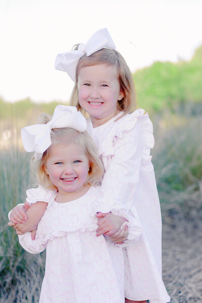 Two young sisters with matching white bows and pastel dresses smile brightly while embracing in a natural outdoor setting. Their joyful expressions and sweet bond capture the warmth and innocence of childhood, reflecting the light and timeless style of a Birmingham family photographer