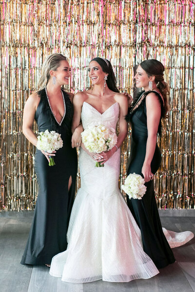 Three women are posing in front of a shimmering, metallic backdrop. The woman in the center is wearing a white wedding gown, holding a bouquet of white flowers, and the two women on either side are in black dresses, also holding white bouquets.