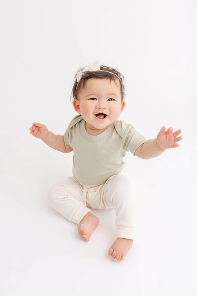 Six Month old baby with dark hair sitting up on her own and smiling at the camera. She is wearing a sage green onesie and cream joggers from Colored Organics and a simple cream bow on her head.