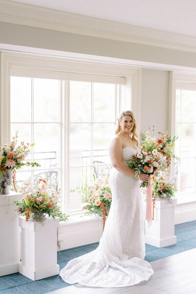 Smiling bride holding peach and white bouquet, standing in front of large windows at Oakville wedding venue