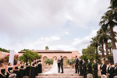 Bride and groom exchange vows during a Royal Palms wedding ceremony, with bridesmaids in black dresses and groomsmen in tuxedos standing on either side under the Arizona sky.